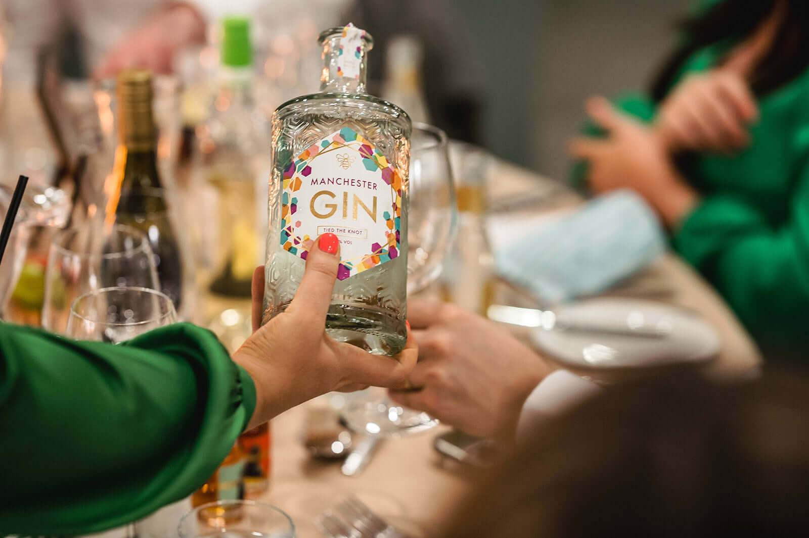 A hand holding a bottle of Manchester Gin at a festive table setting, celebrating love and craft spirits.